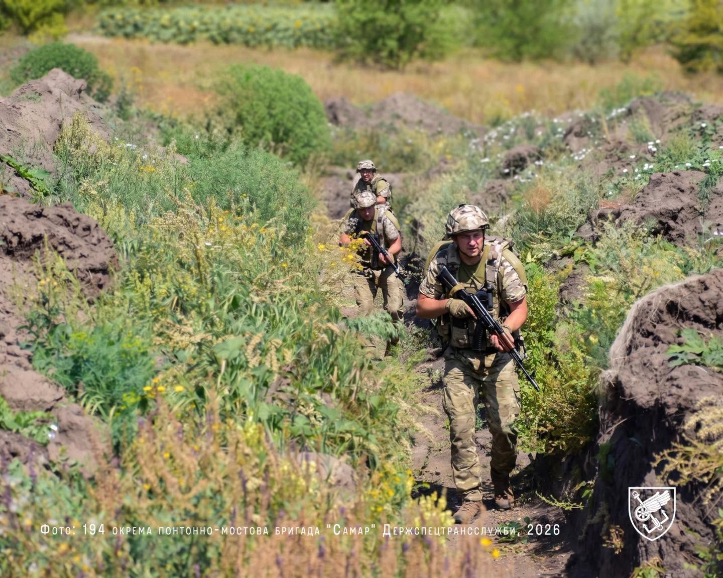 Троє військовослужбовців у камуфляжі та з автоматами рухаються вузькою стежкою серед високої трави й земляних укриттів. Попереду йде боєць, який уважно контролює напрямок руху, тоді як побратими тримають дистанцію позаду.