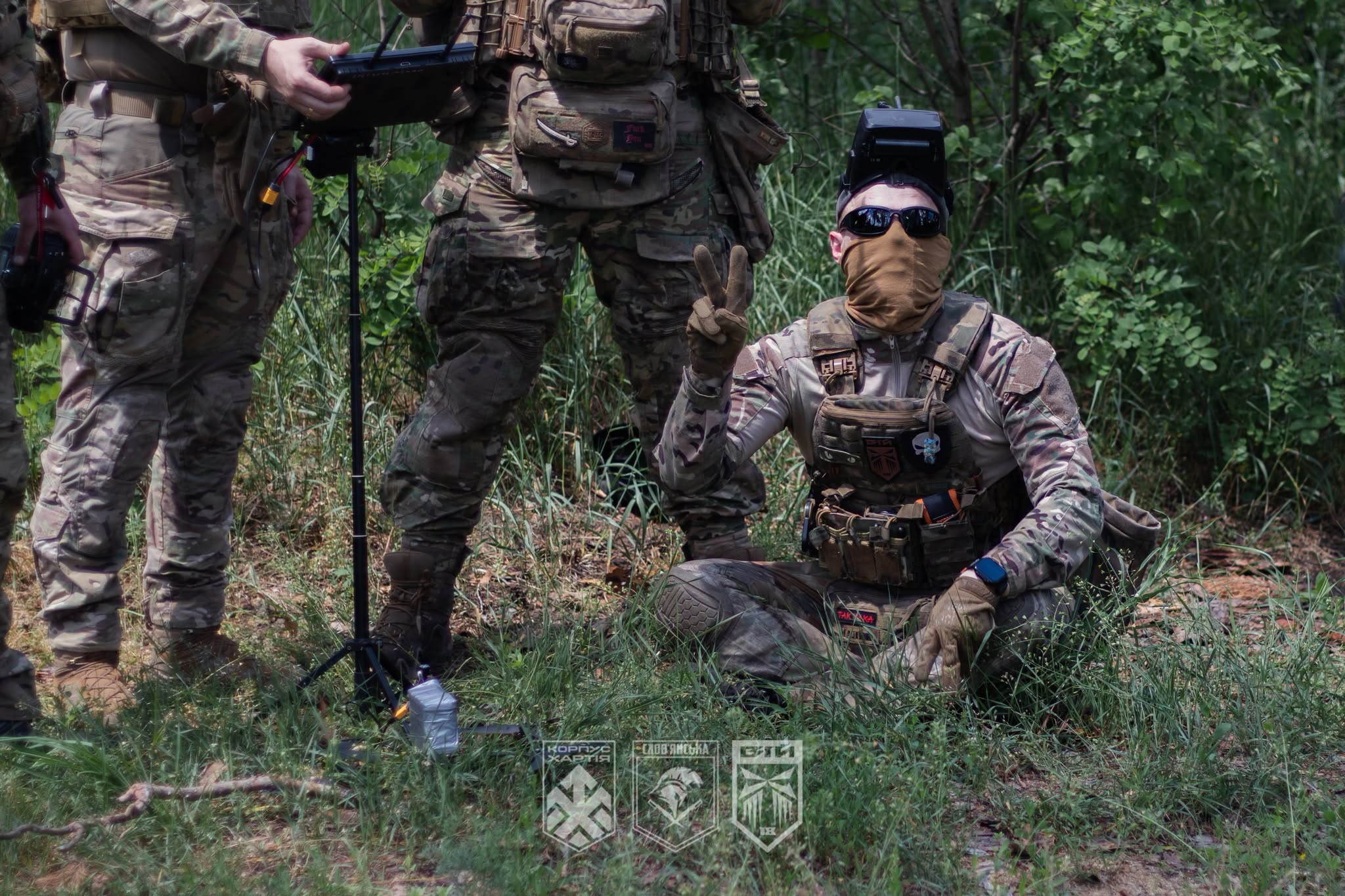 У лісистій місцевості група військовослужбовців у камуфляжі працює з обладнанням для керування безпілотником. Один із бійців сидить на землі в захисних окулярах і масці, показуючи жест перемоги, тоді як його побратими налаштовують апаратуру.