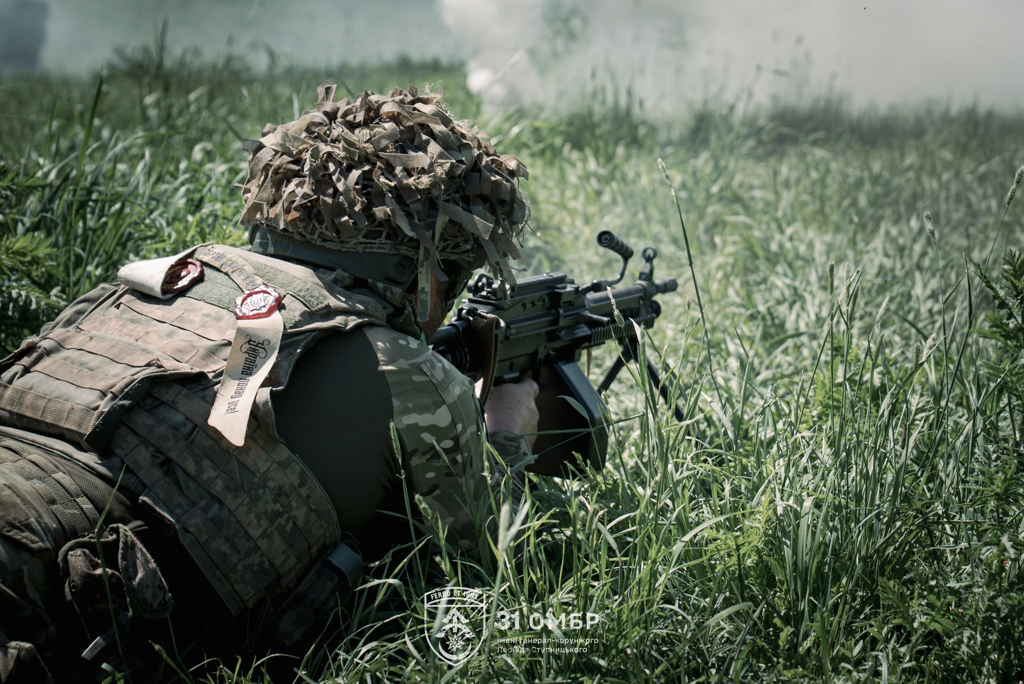 Військовослужбовець у камуфляжі лежить у високій траві та веде прицільне спостереження зі стрілецької зброї. Дим на тлі й низький ракурс передають напружену атмосферу бойової роботи на передовій.