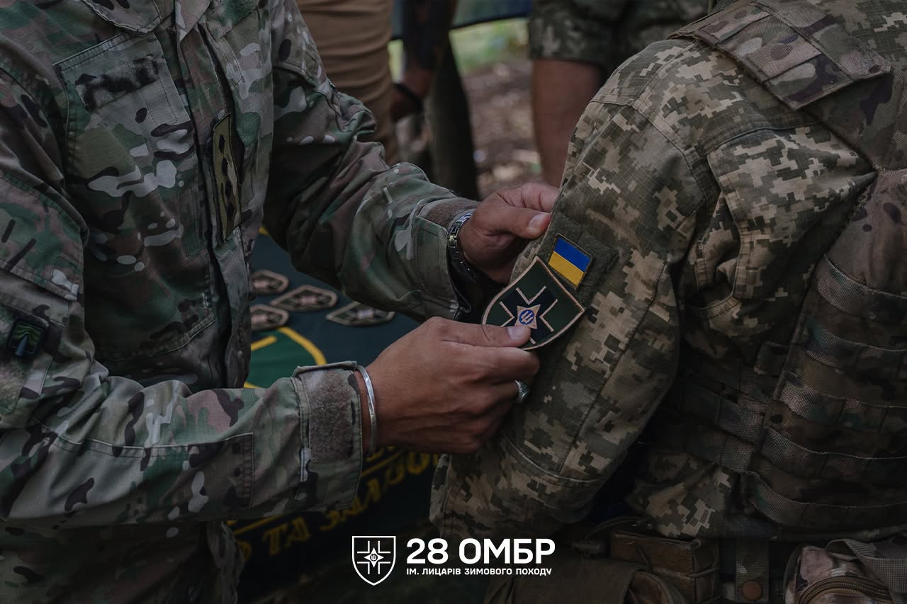 Військовослужбовець прикріплює нарукавний шеврон 28-ї окремої механізованої бригади до форми іншого бійця. Над шевроном розміщений прапор України, а внизу кадру видно символіку бригади імені Лицарів Зимового Походу.