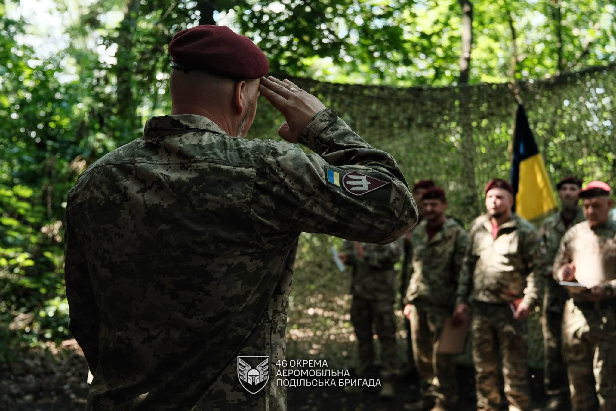 Військовослужбовець у бордовому береті віддає честь перед строєм побратимів у лісистій місцевості. Позаду майорить український прапор, а на формі видно шеврон 46 окремої аеромобільної Подільської бригади.