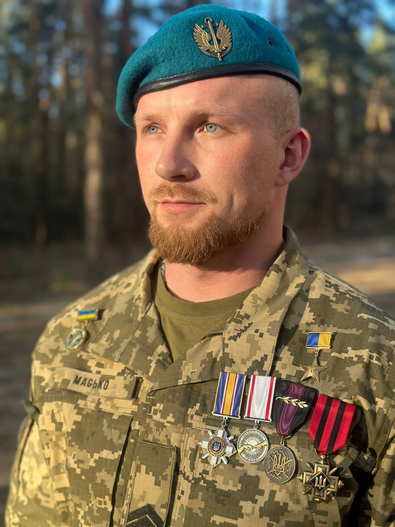 Військовий у камуфляжній формі та блакитному береті дивиться вбік. На його одязі закріплені медалі, нагрудні знаки та нашивка з написом «Масько».