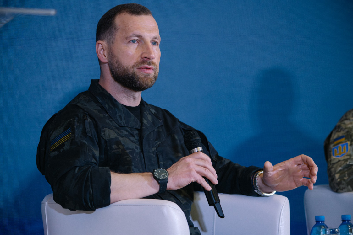 Чоловік у чорній військовій формі сидить у кріслі, тримає мікрофон і жестикулює під час виступу. Позаду нього — великий синій екран, а на рукаві видно нашивку в кольорах українського прапора.