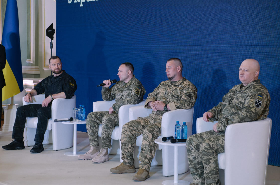 Четверо чоловіків сидять у кріслах на сцені під час панельної дискусії, троє з них одягнені у військову форму. Один із військових говорить у мікрофон, тоді як інші учасники слухають його на тлі синього екрана та українського прапора.