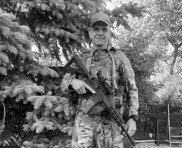 Військовослужбовець у повному спорядженні стоїть просто неба на тлі дерев і тримає перед собою автомат. Чорно-білий кадр передає спокійний момент поза безпосередньою зоною бойових дій.