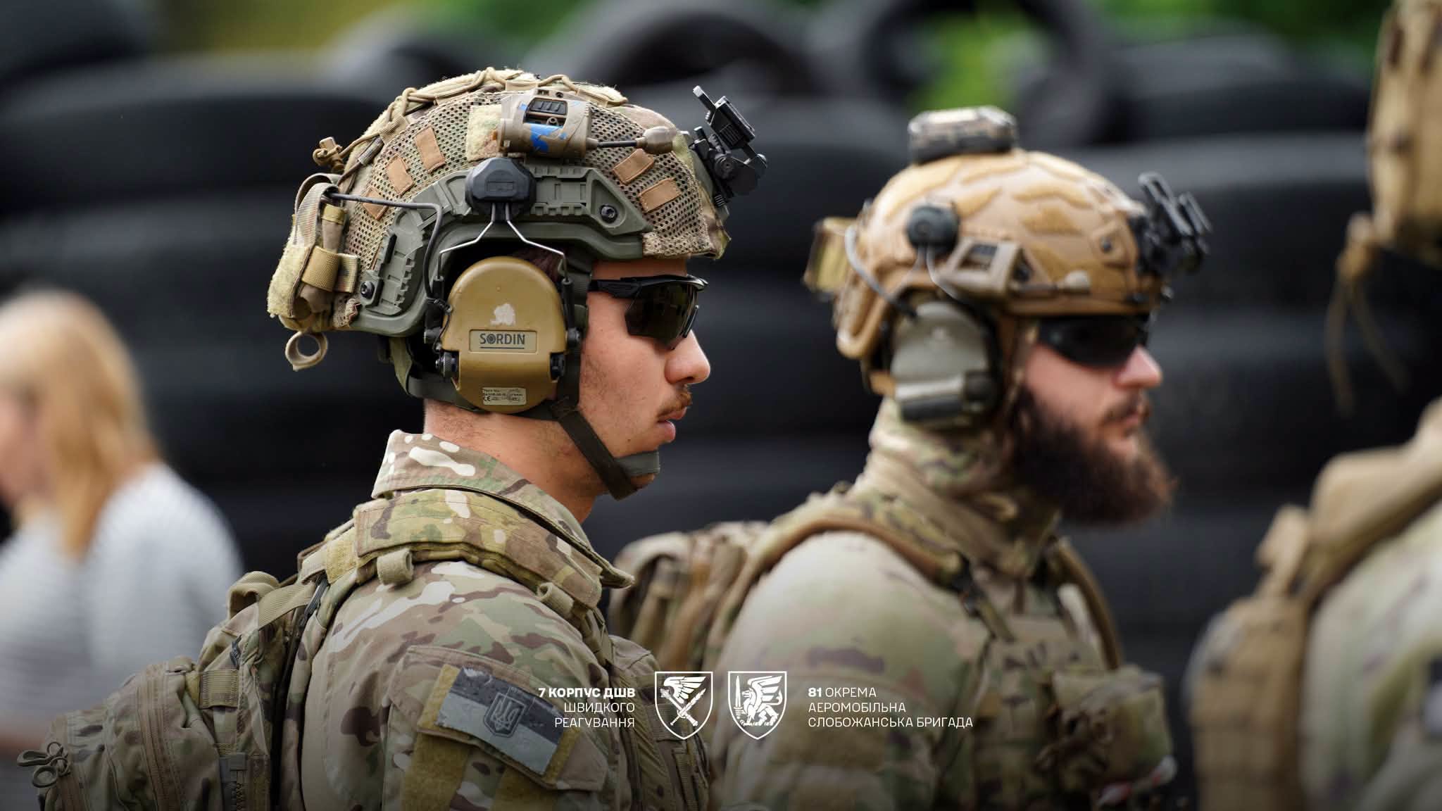 На передньому плані зображений військовослужбовець у шоломі, захисних окулярах і тактичних навушниках. Поруч стоять інші бійці в аналогічному спорядженні, а розмитий фон зосереджує увагу на деталях екіпірування.