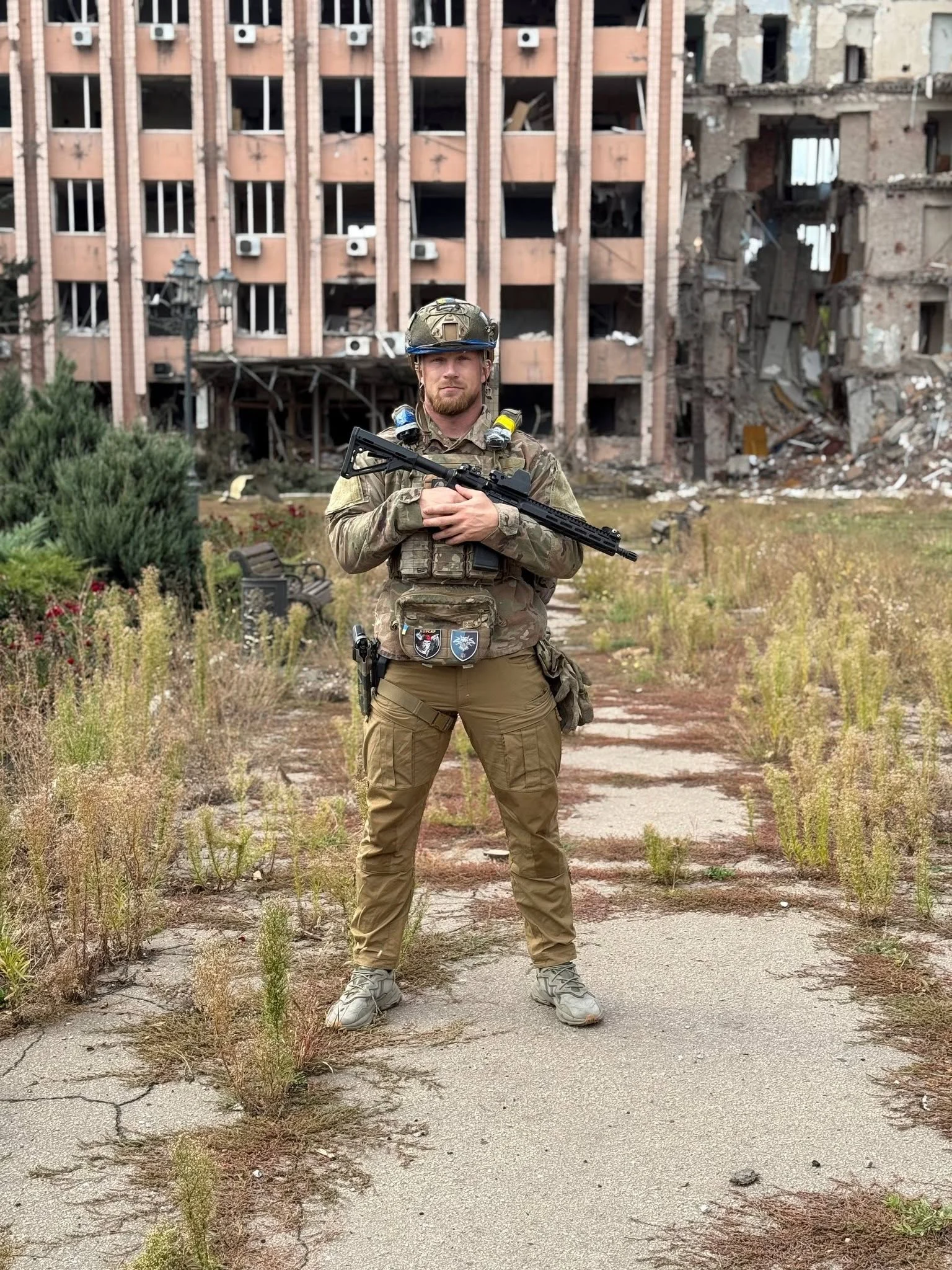 Військовий у спорядженні тримає зброю перед собою. Позаду розташована багатоповерхова будівля з пошкодженими стінами, вибитими вікнами та частково зруйнованими приміщеннями.
