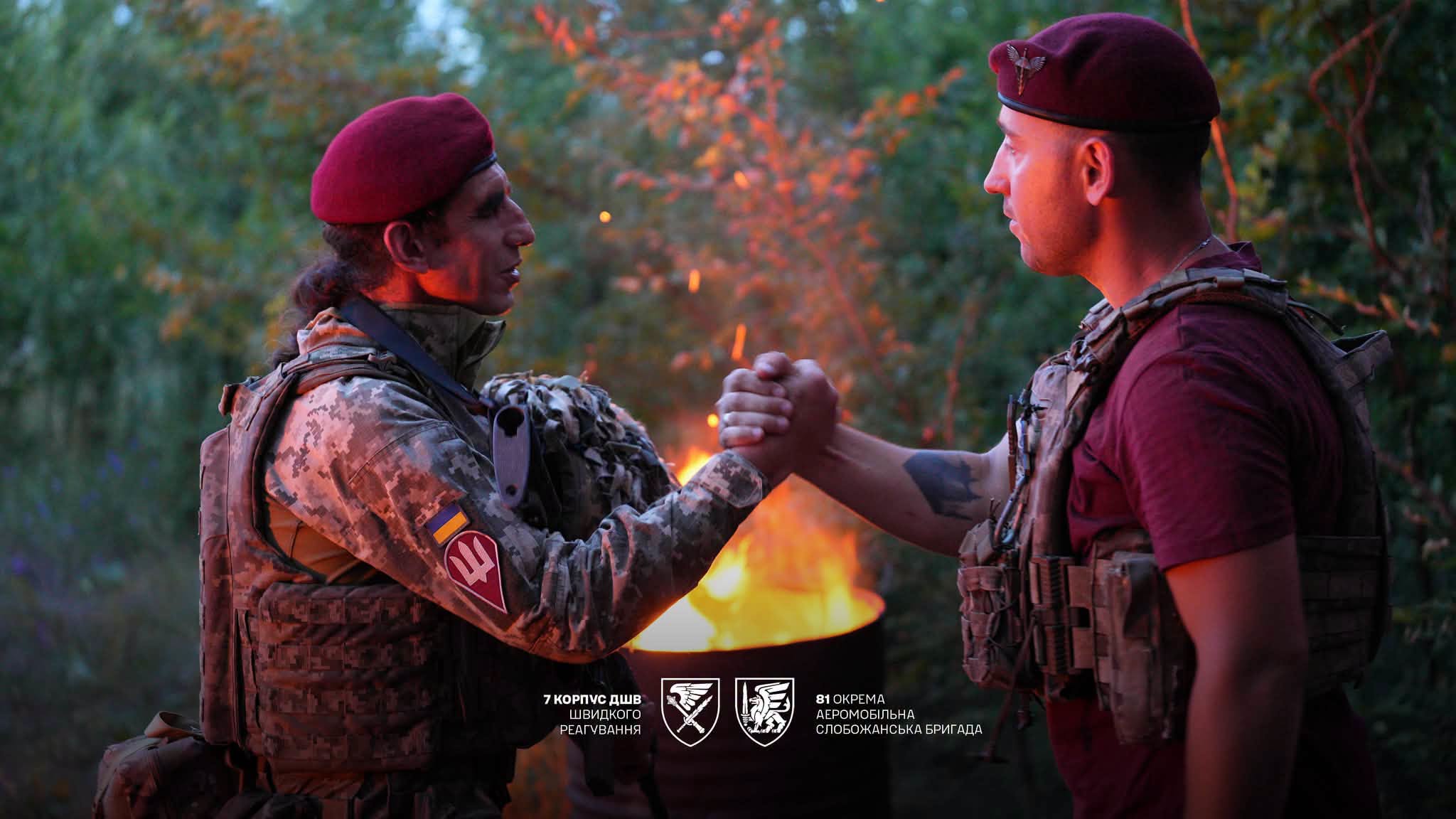 Двоє військовослужбовців у беретах тиснуть один одному руки біля вогнища в лісовій місцевості. Тепле світло полум’я підкреслює урочистий і товариський характер зустрічі.