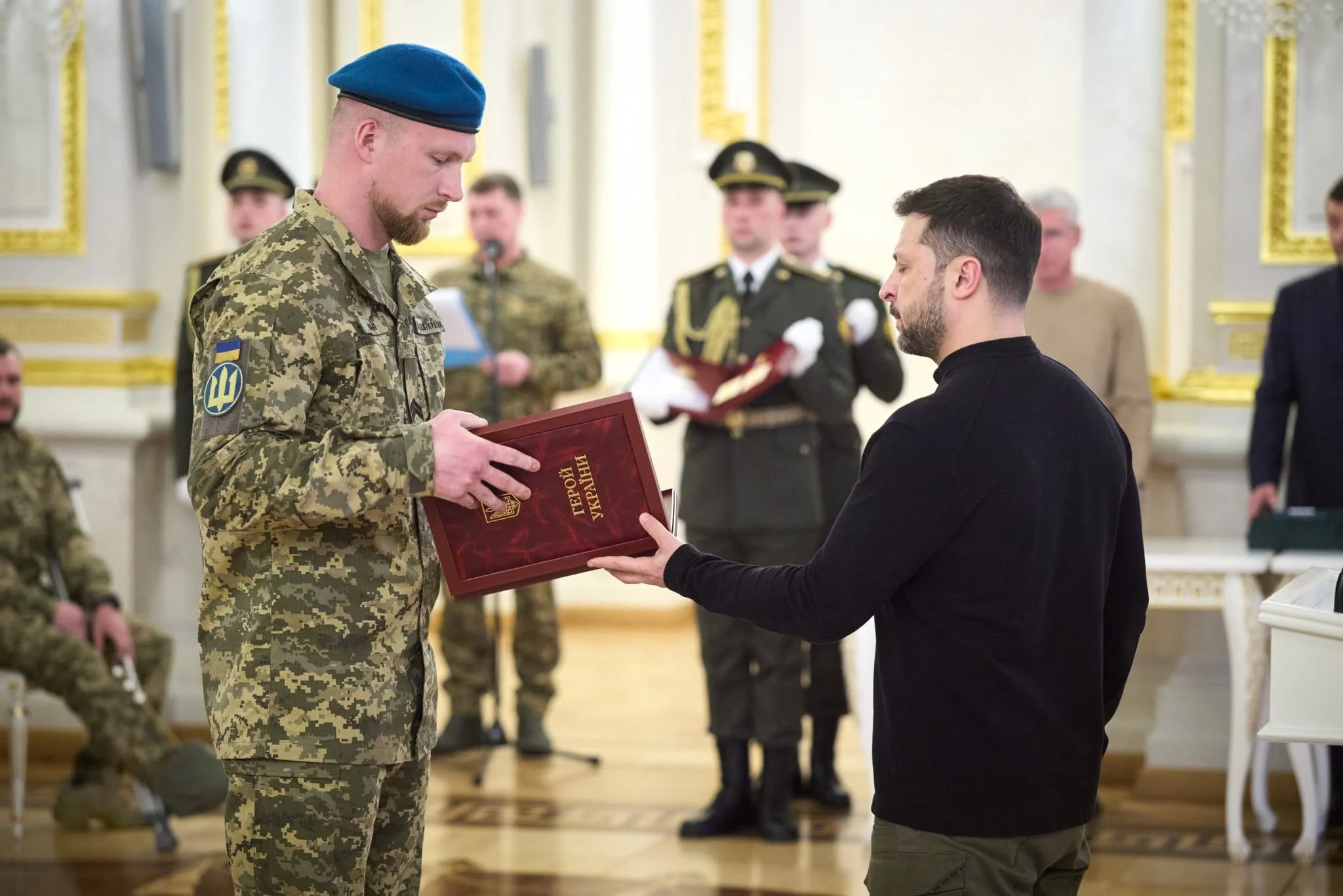 Військовий у блакитному береті та чоловік у чорному светрі тримають червоний футляр із написом «Герой України». Позаду стоять військовослужбовці, зокрема почесна варта з нагородою на подушці.