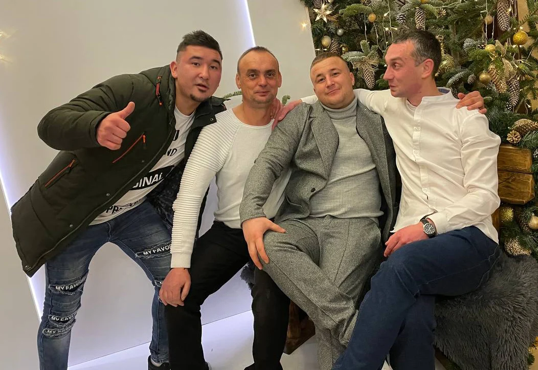Четверо чоловіків сидять поруч і обіймають один одного за плечі. Позаду них стоїть прикрашена ялинка.