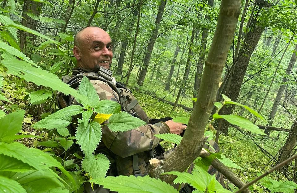 Чоловік у камуфляжному одязі сидить серед дерев і повертає голову вбік. Частину його постаті закривають гілки та листя.