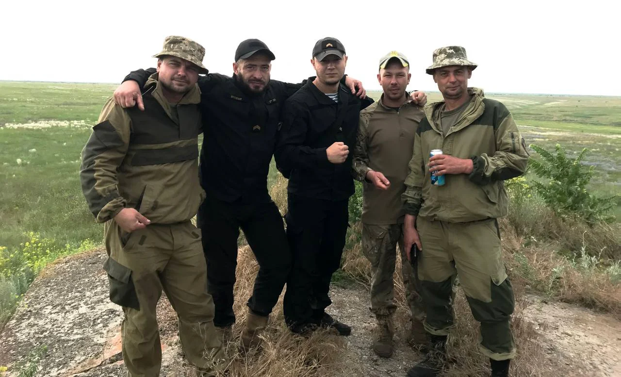 П’ятеро чоловіків стоять разом на відкритій місцевості. Двоє обіймають інших за плечі, а чоловік праворуч тримає бляшанку.